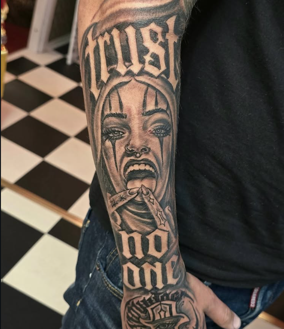 Black and grey underarmstatuering med chicano-lettering "trust no one" och clownflicka